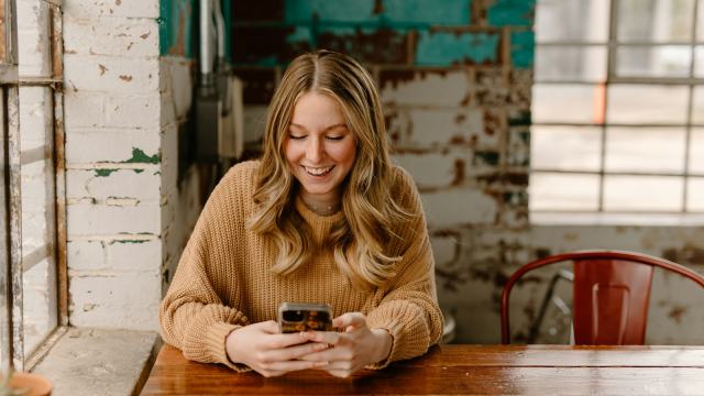 Mulher sorrindo enquanto agenda um horário pelo celular sentada à mesa