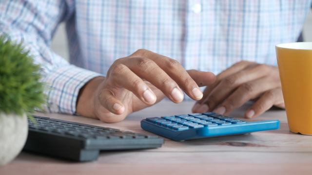 Pessoa fazendo contas em uma calculadora azul ao lado do teclado do computador